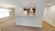 This single-floor residence offers a clean, open-concept design that connects the kitchen and living area in a comfortable, functional layout. The kitchen is outfitted with warm medium-toned wood cabinetry, a white appliance suite including a refrigerator, electric range, and built-in microwave, as well as a dark countertop that provides a subtle contrast to the cabinetry. A peninsula with a light-toned countertop extends into the living space, creating a natural breakfast bar complete with pendant lighting overhead — a practical and stylish divider between the two areas. Recessed lighting supplements the pendant fixtures, ensuring the kitchen remains well-lit. The living area is finished with plush, neutral-toned carpeting that adds warmth underfoot and flows throughout the space. The overall color palette is kept soft and neutral — whites, beiges, and warm wood tones — lending the interior a bright, welcoming ambiance. The layout is thoughtfully designed to maximize usability, making the space well-suited for both everyday living and casual entertaining.