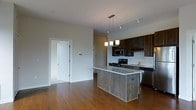 This single-floor space showcases a thoughtfully designed open-concept layout that seamlessly connects the kitchen and living areas. The kitchen is a standout feature, anchored by a substantial island topped with granite and clad in natural wood paneling, providing both functional workspace and a striking visual centerpiece. Above, a modern pendant light fixture casts warm, focused illumination over the island. Dark espresso-toned cabinetry lines the kitchen walls, complemented by a classic white subway tile backsplash that adds texture and contrast. Stainless steel appliances, including a range, microwave, and dishwasher, deliver a sleek, contemporary feel. A stainless steel sink is positioned beneath upper cabinetry with glass-front display panels, adding an elegant touch. Track lighting runs along the ceiling, ensuring the entire space is well-lit. Rich, warm hardwood flooring flows throughout the open area, grounding the space with natural warmth and continuity. The neutral wall tones create a clean, versatile backdrop, while the open layout allows for flexible furniture arrangement. A white interior door leads to an adjacent room, further extending the functional living footprint of this well-appointed space.