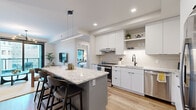 Located on Peachtree Street in Atlanta's vibrant Midtown neighborhood, this thoughtfully designed 2-bedroom, 1-bathroom residence offers a clean, contemporary aesthetic across a well-organized single floor. The open-concept kitchen is a standout, featuring a large granite-topped island with seating, flat-front white cabinetry, open shelving, stainless steel appliances, tile backsplash, and a linear pendant light — all set atop warm hardwood flooring that flows throughout the living areas. The living room is bright and inviting, anchored by floor-to-ceiling windows and a sliding glass door that opens to a private balcony with sweeping views of the Midtown Atlanta skyline and tree-lined streetscape below. The primary bedroom is comfortably sized, accented by a ceiling fan and direct access to the sleek full bathroom, which features a double vanity with illuminated mirrors, marble-look tile, and a glass-enclosed walk-in shower. The second bedroom offers large windows with city views and versatile space ideal for a home office or guest room. Floating wood shelves add functional character. A hallway and dedicated closet complete the efficient layout, making this a polished urban residence with refined finishes throughout.