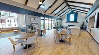 This single-floor community clubhouse is designed to impress with its striking vaulted ceilings adorned with dark exposed wood beams, creating an open and airy atmosphere throughout. Light wood-look flooring flows seamlessly across the main gathering area, which is furnished with multiple round dining tables and modern upholstered chairs, offering ample space for socializing and events. A standout feature is the full-size pool table, positioned near expansive floor-to-ceiling sliding glass doors that open to an outdoor balcony with views of the surrounding neighborhood. The clubhouse includes a well-appointed kitchenette, complete with white cabinetry, a sink, open shelving, and bar-height seating along a large white island. A cozy lounge area is anchored by a sleek, linear fireplace set against a white brick accent wall, complemented by plush seating and patterned carpet. A separate sitting nook features a built-in window bench beneath dramatic floor-to-ceiling windows, bathing the space in natural light. Bold teal accent walls, decorative mirrors, wall-mounted televisions, and carefully chosen lighting fixtures throughout contribute to the space's modern yet inviting character.