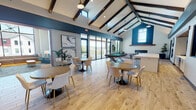 This single-floor community clubhouse is designed to impress with its striking vaulted ceilings adorned with dark exposed wood beams, creating an open and airy atmosphere throughout. Light wood-look flooring flows seamlessly across the main gathering area, which is furnished with multiple round dining tables and modern upholstered chairs, offering ample space for socializing and events. A standout feature is the full-size pool table, positioned near expansive floor-to-ceiling sliding glass doors that open to an outdoor balcony with views of the surrounding neighborhood. The clubhouse includes a well-appointed kitchenette, complete with white cabinetry, a sink, open shelving, and bar-height seating along a large white island. A cozy lounge area is anchored by a sleek, linear fireplace set against a white brick accent wall, complemented by plush seating and patterned carpet. A separate sitting nook features a built-in window bench beneath dramatic floor-to-ceiling windows, bathing the space in natural light. Bold teal accent walls, decorative mirrors, wall-mounted televisions, and carefully chosen lighting fixtures throughout contribute to the space's modern yet inviting character.