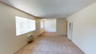 This single-floor unit presents an open-concept living space characterized by neutral, beige-toned walls and wall-to-wall carpeting throughout. The main living area is generously proportioned, offering a comfortable sense of openness and flow. A ceiling fan with a light fixture is centrally positioned, providing both air circulation and ambient lighting. A wall-mounted air conditioning unit is installed beneath the large windows on the left wall, ensuring climate control year-round.

Natural light enters abundantly through a wide window fitted with vertical blinds, brightening the interior and creating an airy atmosphere. The textured popcorn ceiling adds a classic residential finish to the space. Toward the rear, the layout opens into an additional area, possibly a dining space or secondary living zone, visible past a partial wall divider. A doorway leads further into the unit, hinting at additional rooms beyond. A louvered utility panel is mounted on the right wall, adding a functional element to the space.

The overall ambiance is clean, neutral, and move-in ready, with a straightforward layout that offers flexibility for furniture arrangement and personalization.