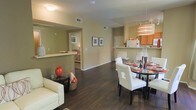 This single-floor residence showcases an open-concept design that integrates the living, dining, and kitchen areas into one cohesive, flowing space. Rich, dark hardwood floors extend throughout, providing a striking contrast against the warm, neutral-toned walls. The living area is furnished with a cream leather sofa and accented with bold red decorative pieces, adding character to the space.

The dining area features a sleek, round glass-top table surrounded by contemporary white upholstered chairs with dark wood legs, positioned beneath a stylish cylindrical pendant light that serves as a focal point. Adjacent to the dining space, the kitchen is accessible via a raised breakfast bar and is equipped with warm wood cabinetry and track lighting overhead.

A hallway visible from the main living area leads to additional rooms, including a bedroom that receives natural light from a window. Framed artwork on the walls and carefully placed decorative accents contribute to a polished, curated aesthetic. The layout maximizes the sense of openness, making the space feel both functional and inviting throughout.