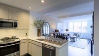 This single-floor residence showcases a thoughtfully designed open-concept layout that seamlessly connects the kitchen and living areas into one cohesive, light-filled space. The kitchen is outfitted with flat-front white cabinetry, a white countertop peninsula with an integrated stainless steel sink, and a stainless steel dishwasher beneath. Recessed ceiling lighting illuminates the crisp, all-white interior, enhancing the clean and contemporary aesthetic throughout. The peninsula serves as both a functional workspace and a social hub, ideal for casual dining or entertaining. A sleek black arched mirror adds a striking decorative accent, complementing the neutral palette. The living area beyond the kitchen benefits from multiple windows that draw in generous natural light, creating a bright and airy atmosphere. Light-toned flooring flows continuously across the space, reinforcing the sense of openness. The living area is furnished with comfortable seating and accent lamps that add warmth to the otherwise minimal design. Together, the kitchen and living spaces form a harmonious, well-appointed interior that balances modern functionality with an inviting residential ambiance.