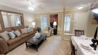 This single-floor living space offers a warm and inviting atmosphere anchored by a thoughtfully designed open layout. The living room serves as the focal point of the home, showcasing elegant crown molding along the ceiling perimeter and a central ceiling fan with integrated lighting. Soft, neutral-toned carpeting runs throughout, complementing the warm beige walls and creating a cohesive, comfortable aesthetic.

Natural light fills the room through multiple windows dressed with classic plantation shutters, offering both brightness and privacy control. A decorative column adds an architectural detail that subtly defines the living area while maintaining an open feel. The space accommodates generous seating arrangements, with room for a sofa, accent chair, and a dark-finished coffee table that grounds the arrangement.

A vibrant abstract painting in warm red and orange tones provides a striking focal point on one wall, adding energy and visual interest to the otherwise neutral palette. Near the entry, a partial glimpse of a dining area suggests a natural flow between living and dining spaces. Overall, the space combines classic architectural details with a relaxed, livable ambiance suitable for everyday comfort.
