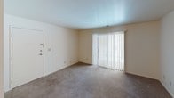 This single-floor, one-bedroom, one-bathroom unit is thoughtfully laid out to maximize everyday functionality. The living area features wall-to-wall carpeting, crisp white walls, and a sliding glass door fitted with vertical blinds that allows natural light to fill the space while providing access to the outdoors. The entry door opens directly into this main living area, creating a straightforward and practical flow throughout the unit. The bedroom is a comfortable, carpeted room with a window fitted with horizontal blinds, offering a pleasant view of surrounding greenery and bringing in additional natural light. A closet is conveniently located within the bedroom. The kitchen is equipped with rich, dark wood cabinetry providing ample storage, a speckled laminate countertop, a stainless steel single-basin sink, and a white refrigerator. Under-cabinet lighting adds a functional touch to the workspace. The full bathroom features wood-toned cabinetry with a white countertop vanity, a large mirror with a multi-bulb vanity light fixture above, a toilet, and a full-size soaking tub with a shower combo. Wood-look flooring in the bathroom adds a warm contrast to the neutral tones found throughout the rest of the unit. Overall, the space offers a clean, neutral palette well-suited for a variety of living arrangements.
