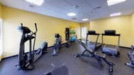 Located at 25 Forest Acres in Haverhill, Massachusetts, this fitness center is a well-equipped, single-floor exercise facility designed to support a comprehensive workout experience. The room features warm yellow walls and a drop-tile ceiling with recessed panel lighting that ensures consistent, bright illumination throughout the space. Durable rubber flooring covers the entire area, providing both safety and comfort during exercise.

The cardio section is equipped with multiple treadmills, elliptical trainers, and a stationary cycle, offering a variety of aerobic workout options. A dedicated strength training area includes a multi-function cable machine, an adjustable weight bench, and a full rack of dumbbells spanning a wide range of weights. A wall-mounted flat-screen television is positioned for easy viewing during workouts.

Complementing the equipment is a ball rack holding several exercise balls in varying sizes, along with a wall-mounted mat storage rack, making the space suitable for stretching and functional fitness routines. A water cooler and a stainless steel waste receptacle are conveniently placed within the room. Natural light is introduced through a window with blinds, and a clearly marked exit door ensures safe egress. Overall, the space is thoughtfully organized to accommodate both cardio and strength training in a clean, functional environment.