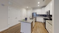 This single-story residence is designed with a clean, modern aesthetic and a practical open-concept layout. The kitchen serves as a central highlight, featuring white shaker-style cabinetry, granite countertops, a dark subway tile backsplash, and a full suite of stainless steel appliances including a refrigerator, range, and microwave. A kitchen island with an integrated sink and matching granite surface provides additional workspace and storage. Wood-style plank flooring runs throughout the main living and kitchen areas, adding warmth and continuity.

The living area is bright and open, accented by crown molding and a ceiling fan, with French doors that invite natural light. Bedrooms are finished with plush carpet and ceiling fans, offering a comfortable, quieter environment. At least one bedroom includes a walk-in closet with wire shelving for organized storage.

Two full bathrooms are present in the unit. One features a soaking tub and shower combination with white subway tile surround, a granite-topped white vanity, and wood-style flooring. The second bathroom offers a similar vanity finish and a standalone shower, maintaining a consistent design throughout. The overall palette of neutral tones and white trim creates a cohesive, light-filled atmosphere.