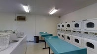 This single-floor laundry facility is designed with functionality and efficiency in mind. The room features white walls and a light-colored floor, creating a clean and bright environment enhanced by fluorescent lighting mounted to the ceiling. Multiple stacked washer and dryer units line the right wall, providing ample laundry capacity for shared or communal use. The machines are arranged in a stacked configuration, maximizing the use of vertical space and allowing for a greater number of units within the room's footprint. Two teal-colored folding tables are centrally positioned, offering convenient surfaces for sorting and folding laundry. A utility sink is visible along the left wall, adding further practicality to the space. A wall-mounted air conditioning unit ensures comfortable conditions year-round. A waste bin is conveniently placed near the folding tables. Overall, the space is straightforward and utilitarian, prioritizing ease of use and practicality, making it well-suited as a shared laundry facility.