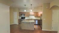 This single-floor space centers around a well-appointed open kitchen that seamlessly integrates with the surrounding living area. The kitchen is equipped with stainless steel appliances, including a refrigerator, dishwasher, electric range, and built-in microwave. Warm-toned wooden cabinetry lines the walls, offering generous storage, while a tiled backsplash adds a clean, polished finish. The countertops extend to a curved central island, which provides additional prep space and accommodates seating, making it ideal for casual dining or entertaining. A double sink is positioned along the perimeter counter beneath additional cabinetry. Lighting is thoughtfully layered, combining two elegant pendant lights over the island with a track lighting system along the ceiling to illuminate the full kitchen workspace. Hardwood flooring runs throughout, contributing warmth and continuity to the space. Neutral yellow-toned walls create a bright, welcoming atmosphere. A white paneled door is visible near the kitchen, suggesting access to a utility or storage area. Overall, the space offers a functional and inviting layout suited for everyday living.
