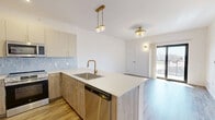 Located on E Second St in Dayton, Ohio, this modern one-bedroom, one-bathroom residence offers a thoughtfully designed open-concept layout with contemporary finishes throughout. Light-toned hardwood-style flooring flows seamlessly across the entire unit, complementing the clean white walls and bright, airy ambiance.

The combined kitchen and living area serves as the heart of the home. The kitchen features a peninsula island with a quartz countertop, a stainless steel undermount sink with a brushed gold faucet, wood-grain lower cabinetry with gold hardware, and a light blue tile backsplash. A full suite of stainless steel appliances includes a refrigerator, range, microwave, and dishwasher. Stylish pendant and flush-mount gold light fixtures add a refined touch overhead.

The spacious primary bedroom is bathed in natural light from a large window and features built-in wood-grain cabinetry, a ceiling fan, and access to multiple closets. Sliding glass doors lead to a private balcony offering open views of the Dayton skyline.

The bathroom is elegantly appointed with large-format marble-look tile flooring, a quartz vanity with wood cabinetry and gold hardware, a backlit mirror, and a glass-enclosed shower with white subway tile. Three closets provide generous storage throughout the residence.