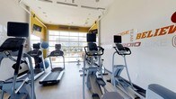 This fitness center is designed to provide a functional and energizing workout environment. The space is anchored by a striking large-format garage-style glass door along the far wall, allowing abundant natural light to pour in while offering views of the surrounding exterior. This architectural feature creates an open, airy atmosphere that enhances the overall workout experience.

The room is equipped with a lineup of modern cardio machines, including treadmills, ellipticals, and stationary bikes, each outfitted with individual display screens for entertainment or performance tracking. The equipment is thoughtfully arranged to maximize floor space and allow comfortable movement throughout the room.

The interior design features a clean white ceiling with recessed lighting supplemented by natural light. Bold yellow accent walls frame the garage door, adding a vibrant, energetic character to the space. The right-side wall features large motivational typography in a mix of red and grey tones, with words such as "TEAM," "CARDIO," "PUSH," and "HEALTH," reinforcing an active and motivating atmosphere. The flooring is a light wood-toned surface, providing a polished, contemporary finish that complements the overall design.