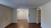 This single-floor residence presents a straightforward and functional layout designed for comfortable everyday living. The interior is characterized by rich, dark hardwood-style flooring that runs consistently throughout the main living areas, providing a warm and cohesive foundation for the space. Neutral white walls create a bright, clean backdrop that allows for versatile décor and furnishing options.

The open layout connects the main living area to an adjoining room, offering a sense of flow and continuity throughout the floor plan. A ceiling fan with a built-in light fixture is installed in the secondary room, contributing both to air circulation and ambient lighting. The textured white ceiling adds a subtle architectural detail across the space.

To one side, a built-in shelving or storage unit is visible, offering practical organizational space integrated directly into the wall. A wall-mounted HVAC vent is positioned overhead, indicating a central climate control system. The overall ambiance of the space is clean, minimal, and move-in ready, with a neutral palette that suits a wide range of interior styles. The open, uncluttered nature of the layout makes the space feel open and adaptable to various living arrangements.