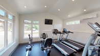 This fitness center in Marlborough, MO is a thoughtfully designed, single-floor facility built to support a well-rounded workout routine. The space is divided into distinct functional zones, each outfitted with dedicated equipment to accommodate a variety of exercise preferences. The cardio area features multiple Spirit treadmills, a recumbent bike, elliptical trainers, and a rowing machine, all arranged to take full advantage of the room's natural light. Large windows along the front wall invite views of the surrounding greenery, while clerestory windows along the side walls further brighten the space. Vaulted ceilings in the cardio zone add an open, airy quality, and recessed lighting ensures consistent illumination throughout. A wall-mounted flat-screen television is positioned for easy viewing during cardio sessions. The strength training area is lined with floor-to-ceiling mirrors that expand the sense of space and assist with form. It includes a multi-functional cable machine, a dumbbell rack with a full range of free weights, and a flat bench, with ample open floor space for stretching or mat work — a pink yoga mat is on hand for this purpose. Durable rubber flooring throughout provides both safety and comfort. The facility is climate-controlled with a wall-mounted mini-split unit and is accessible via a dedicated exterior entrance.