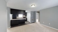 This single-floor residence at The Fields offers a thoughtfully designed open-concept layout that connects the kitchen and living areas in a cohesive, functional arrangement. The kitchen is appointed with bold dark cabinetry that reaches toward the ceiling, creating a striking contrast against the light grey walls. Black appliances, including a refrigerator and range, complement the cabinetry and contribute to the space's contemporary aesthetic. A window above the sink introduces natural light, brightening the workspace. Dark hardwood-style flooring anchors the kitchen zone, while plush neutral carpet defines the living area, providing a comfortable and warm transition between spaces. The grey walls throughout lend a modern, neutral palette that pairs well with the darker design elements. A closet with a classic white paneled door adds convenient storage, while a hallway leads to additional rooms beyond, suggesting a practical and well-organized floor plan. A ceiling-mounted light fixture provides central illumination, and a floor vent ensures efficient climate control. The overall ambiance is clean, modern, and move-in ready, with a balanced combination of dark and light tones throughout.