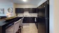 This single-floor, two-bedroom, one-bathroom residence provides a thoughtfully laid-out living environment with functional design throughout. The kitchen is a standout feature, equipped with dark espresso cabinetry, light countertops, black appliances including a range, microwave, and refrigerator, and durable wood-look flooring. A dining area adjacent to the kitchen accommodates a bar-height table with upholstered seating, making it ideal for casual meals and entertaining. The living room offers a welcoming atmosphere with neutral-toned walls, wood-look flooring, and ample space for comfortable seating arrangements. The primary bedroom features plush carpeting, a ceiling fan, and warm ambient lighting, creating a restful retreat. The second bedroom is equally versatile, also carpeted and ceiling fan-equipped, with a window that allows natural light to fill the room and includes a closet for storage. The shared full bathroom is cleanly finished with a dark vanity cabinet, a large mirror, tile flooring, and a shower-tub combination. Throughout the residence, neutral wall tones and consistent finishes create a cohesive and modern aesthetic, making this a well-appointed and practical living space.