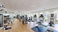 Located at Danville Park Drive SW in Decatur, Alabama, this fitness center offers a spacious, single-floor layout designed to accommodate a comprehensive range of workout activities. The open floor plan is finished with smooth hardwood-style flooring throughout, providing a clean and functional surface ideal for both equipment use and mat-based exercises.

The facility is outfitted with a robust selection of cardio equipment, including multiple treadmills, elliptical trainers, stationary bikes, and a recumbent cycle. A dedicated strength training area features several multi-function cable and weight machines suited for targeting various muscle groups, complemented by a free weights station with a full dumbbell rack. Exercise mats are positioned throughout for stretching and floor workouts.

Large windows along the exterior walls allow ample natural light to fill the space while offering views of the surrounding greenery, creating an energizing atmosphere. Full-length mirrors line multiple walls, visually expanding the room and providing functional feedback during workouts. Recessed lighting and a drop ceiling with integrated ventilation ensure a comfortable indoor environment year-round.

Motivational wall graphics and a wall-mounted flat-screen television add to the engaging ambiance. A clearly posted fitness center rules board is visible near the entrance, and access is provided through glass-paned double doors. The overall design is clean, modern, and well-maintained, making it a fully functional community fitness facility.