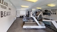 This single-floor fitness room offers a practical and functional workout environment designed to accommodate a range of exercise needs. The space features a textured ceiling and grey-painted walls, with flush-mounted fluorescent light fixtures providing consistent, even illumination throughout. A window fitted with vertical blinds allows natural light to filter in, contributing to an open and energizing atmosphere.

The room is equipped with multiple cardio machines, including a treadmill, elliptical trainer, and stationary bike, arranged to maximize the available floor space. A cable weight station positioned along one wall supports strength training exercises, while stability balls stored on dedicated stands offer flexibility for core and balance workouts. The dark, durable flooring is well-suited for high-traffic fitness use.

A large wall-mounted mirror runs along one side of the room, visually expanding the space and allowing users to monitor their form during exercise. A wall-mounted television adds an entertainment option for those engaging in longer cardio sessions. A motivational wall decal reading "Release Your Inner Beast" adds a spirited, energetic character to the room. Entry is provided through a solid door, and the overall layout is efficiently organized to support a comprehensive fitness routine.