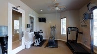 This single-floor fitness room offers a focused and functional workout environment designed to support an active lifestyle. The space is outfitted with a selection of cardio equipment, including a treadmill, an elliptical trainer, and a stationary exercise bike, all positioned on protective floor mats for safety and stability. A wall-mounted flat-screen television is installed at eye level, providing entertainment during workouts. The room is kept comfortable by a ceiling fan with an integrated light fixture, which promotes air circulation throughout the space. Natural light enters through a window fitted with horizontal blinds, adding brightness to the neutral, light-toned walls and enhancing the overall ambiance. Recessed lighting supplements the room's illumination. An interior glass-paneled door connects the fitness room to an adjacent area, contributing to an open and accessible feel. The clean, minimalist design of the space keeps the focus on functionality, making it a practical and welcoming environment for daily exercise routines.