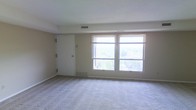 This single-floor space presents a clean, open room finished with wall-to-wall carpeting in a neutral beige tone, contributing to a quiet and understated aesthetic. The walls are painted in a soft, warm neutral shade that complements the light palette throughout. Two large windows dominate one wall, fitted with horizontal blinds and allowing an abundance of natural light to fill the room. The windows offer elevated views of surrounding trees and the broader outdoor environment, adding a pleasant sense of openness. A white door is positioned along the adjacent wall, providing access to another area of the space. Overhead, a textured ceiling adds subtle visual interest, while ceiling vents indicate a central HVAC system for climate control. A baseboard heater is also present beneath the windows, offering supplemental warmth. The overall ambiance is bright, airy, and move-in ready, with a simple and functional layout that provides flexibility for a variety of uses.