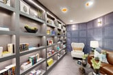 Located on District Lane in Chattanooga, Tennessee, this thoughtfully designed library lounge offers a refined and inviting atmosphere ideal for reading and relaxation. The space is anchored by a dramatic floor-to-ceiling built-in shelving unit finished in a sleek gray tone, housing an extensive collection of books alongside decorative objects, potted succulents, and sculptural accents. Integrated LED strip lighting illuminates each shelf, adding warmth and visual depth to the display. The opposite corner features a striking blue-gray embossed wallcovering with a geometric panel pattern that serves as a bold focal point, flanked by stylish wall sconces that cast a soft ambient glow. Seating is both varied and comfortable, with a plush white wingback chair, a rich cognac leather sofa adorned with teal accent pillows, and round metallic side tables that complete the lounge arrangement. The flooring is finished in a light herringbone-patterned wood-look material, adding texture and an understated elegance underfoot. Recessed ceiling lighting provides even illumination throughout, complementing the layered lighting scheme. The overall ambiance is one of sophisticated comfort — a quiet, curated retreat that balances modern interior design with a warm, literary character.