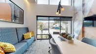 This contemporary single-floor office space is thoughtfully designed to balance functionality with modern aesthetics. The interior is immediately distinguished by its soaring double-height ceilings, which flood the space with natural light and create an open, airy atmosphere. Large floor-to-ceiling windows with black metal framing offer direct views of the surrounding streetscape and greenery, seamlessly connecting the interior to the outside environment.

The main workspace features a long, light-wood desk paired with sleek gray ergonomic chairs, ideal for focused work or collaborative sessions. A vibrant blue sofa accented with yellow and orange throw pillows adds a pop of color and provides a comfortable lounge area. A geometric-patterned area rug anchors the space with texture and visual interest.

A dedicated conference room is a standout feature, boasting a dramatic geometric mural wall in neutral tones of gray, beige, and white. The room is equipped with a wall-mounted display screen and a central conference table surrounded by upholstered chairs — well-suited for meetings and presentations.

Connecting hallways are finished with textured carpet, elegant wall sconces, and glass-paneled interior doors that maintain visual openness. Built-in cabinetry with a dark stone countertop and curated sculptural décor adds refined storage and artistic detail. Throughout, the space strikes a balance of professional efficiency and inspired design.