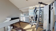 The Upstairs Fitness Center is a light-filled exercise space designed to support a well-rounded workout routine. Situated on an upper level accessible by a carpeted staircase with a white-railed balustrade, the room features soaring vaulted ceilings and an expansive bank of large windows that flood the space with natural light and offer pleasant views of surrounding trees and greenery. The open floor plan is carpeted throughout and provides ample room to move between equipment without feeling cramped. Strength training is well-represented with a multi-station cable machine that accommodates a variety of exercises, a set of mirrors along one wall to assist with form, and a full rack of dumbbells ranging across multiple weight increments. Cardiovascular options include stationary upright and recumbent exercise bikes, catering to users of varying fitness levels and preferences. A flat workout bench is also available for free weight exercises. The bright, airy atmosphere, combined with the natural light streaming in through the generous windows, creates an energizing and comfortable environment for exercise. Fitness center rules are posted on-site for user reference, ensuring a well-managed and accessible facility for all members.