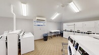 Located at 2500 Knights Rd in Bensalem, PA, this community laundry facility offers a well-organized, single-floor layout designed to accommodate high-volume laundry needs efficiently. The space is finished with neutral-toned square floor tiles and bright white walls, creating a clean and practical environment throughout.

The room is generously outfitted with a large bank of stacked dryers lining one wall, providing substantial drying capacity. Complementing these are multiple top-load and front-load washing machines arranged in rows, offering users flexible options for their laundry needs. A wall-mounted mini-split HVAC unit ensures year-round climate comfort within the space.

Overhead, a combination of recessed skylights and fluorescent strip lighting floods the room with ample illumination, enhancing the bright and airy atmosphere. Ceiling fans further contribute to air circulation. Folding tables and laundry carts are conveniently available for resident use, while informational signage is posted on the walls for easy reference.

The facility is accessed through a set of glass doors that allow natural light to enter from the exterior, adding to the open feel of the space. A fire extinguisher and emergency exit signage reflect a commitment to safety standards. Overall, this laundry facility is thoughtfully designed to be functional, accessible, and well-maintained.