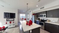 This contemporary single-floor residence showcases a sleek, modern design with an open-concept layout that seamlessly connects the kitchen, dining, and living areas. The kitchen is a standout feature, anchored by a large island with a light quartz countertop, undermount sink, and stylish pendant track lighting overhead. Dark espresso cabinetry contrasts sharply with white countertops, while stainless steel appliances — including a French door refrigerator, range, and microwave — complete the space. The living room is bright and airy, benefiting from a generous bank of windows that invite natural light and provide views of the surrounding outdoor areas. Wood-look flooring runs throughout the main living spaces, adding warmth to the clean aesthetic. The primary bedroom offers plush carpeting, a ceiling fan, walk-in closet, and direct access to a private outdoor area. Its en-suite bathroom features a dark vanity with quartz countertop and a combined tub and shower enclosure with dark tile surround. A second bedroom mirrors similar design qualities, also with outdoor access. A compact half bath with wood-look flooring and framed artwork rounds out the space. Throughout, the residence maintains a cohesive palette of white, dark wood, and stainless steel accents for a polished, contemporary feel.
