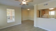 This single-floor space showcases an open-concept layout that seamlessly connects the living, dining, and kitchen areas. The main living area is finished with smooth hardwood-style flooring throughout, lending a clean and cohesive look underfoot. Crown molding lines the ceiling, adding a touch of architectural refinement to the otherwise understated design. A ceiling fan with a built-in light fixture is centrally mounted, providing both functionality and a focal point overhead.

The living and dining areas flow naturally into one another, separated subtly by a column detail that adds structural character without interrupting the openness of the space. Neutral-toned walls complement the light flooring, creating a bright and airy atmosphere. A window fitted with blinds allows natural light to filter in from one side of the room.

Adjacent to the main living area, a pass-through counter connects to the kitchen, which features warm wood-toned cabinetry. This passthrough design enhances the sense of connectivity between spaces while maintaining a defined kitchen zone. Overall, the space presents a functional and well-detailed interior with classic finishing touches.