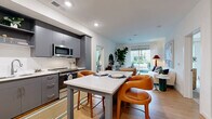 Unit 431 at 111 E Broad St in Falls Church, Virginia offers a thoughtfully designed two-bedroom, two-bathroom residence on a single floor. The open-concept living and dining area is anchored by light hardwood flooring and large sliding glass doors that lead to a private balcony, flooding the space with natural light and providing outdoor views. The kitchen features sleek gray cabinetry, stainless steel appliances, white quartz countertops, and a central island with bar seating — ideal for both cooking and casual dining. The primary bedroom is a comfortable retreat with a walk-in closet outfitted with wire shelving, and a large window framing greenery outside. An accent wall with floral wallpaper adds a distinctive touch. The second bedroom is equally inviting, featuring natural light from a generous window. Both bathrooms are finished with classic black-and-white basketweave tile flooring and dark gray vanities with white countertops. One bathroom includes a tiled walk-in shower, while the other features a full soaking tub. Additional storage is provided by three closets and a utility room, rounding out this well-appointed Falls Church residence.