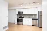 This single-floor unit offers a clean, contemporary layout designed for efficient and comfortable living. The kitchen is a standout feature, outfitted with a full suite of stainless steel appliances — including a range, microwave, dishwasher, and refrigerator — complemented by crisp white cabinetry, a textured tile backsplash, and track lighting that illuminates the workspace effectively. The open design allows the kitchen to flow naturally into the main living area, which is finished with warm hardwood-style flooring that extends throughout the unit, creating a cohesive and inviting atmosphere.

The living space benefits from generous natural light, with large floor-to-ceiling sliding glass doors opening to an outdoor balcony, offering views of the surrounding neighborhood and greenery. Neutral white walls and clean lines throughout contribute to a bright, airy feel. Additional rooms provide versatile spaces suitable for a bedroom or home office, each featuring the same hardwood-style flooring, floor vents for climate control, and access to natural light.

The bathroom is well-appointed with a wide vanity, an undermount sink, and a large mirror. A practical stacked washer and dryer unit is conveniently located within the bathroom, providing in-unit laundry — a notable functional feature. Overall, the space presents a modern, move-in-ready environment with thoughtful finishes.