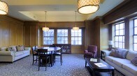 Located at 1301 Washington St in Vicksburg, Mississippi, this distinguished single-floor space exudes a timeless, refined character. Rich wood paneling lines the walls from floor to crown molding, lending a sense of traditional craftsmanship and warmth throughout. Large multi-pane windows allow generous natural light to pour in, framing views of the surrounding greenery and creating a bright, welcoming atmosphere. The interior is anchored by an ornate patterned carpet in soft blue-grey tones, which complements the deep, dark wood furnishings found throughout. A round dining or meeting table surrounded by dark wooden chairs serves as a central gathering point, while plush upholstered seating — including a rich purple armchair — offers comfortable lounging areas. Elegant pendant light fixtures with decorative shades hang from the ceiling, adding a sophisticated, layered lighting scheme that enhances the room's classic ambiance. Additional accent furniture, including side tables and a display cabinet, contribute to the space's polished, well-appointed feel. The overall atmosphere is one of understated elegance, ideal for both formal gatherings and relaxed conversation in a historically rich setting.