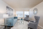 This single-floor residential unit offers a thoughtfully arranged layout that balances functionality and comfort throughout its key living spaces. The galley-style kitchen is outfitted with white cabinetry featuring brushed nickel hardware, light countertops, a double sink, and white appliances including a refrigerator, electric range, and microwave. Wood-look flooring runs through the kitchen and flows into the adjacent dining area, which is illuminated by a pendant light fixture and comfortably accommodates a round glass dining table with tufted upholstered chairs. The living room provides a welcoming retreat with large windows dressed in horizontal blinds that invite natural light and outdoor views. Hard surface flooring anchors the space, complemented by a textured ceiling overhead. Two bedrooms feature plush carpet flooring, generous windows, and textured ceilings, offering a quiet and restful atmosphere. The bathroom is finished with white tile surrounding a full bathtub and shower combination, complete with a safety grab bar, alongside standard fixtures in a clean, neutral palette. Throughout the unit, a cohesive neutral color scheme provides a versatile backdrop, while consistent design details tie each room together into a comfortable and practical living environment.