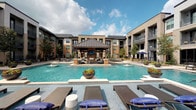 Located in Stafford, Texas, this single-floor residence is part of a contemporary multi-story community designed with upscale living in mind. The exterior courtyard is a standout feature, showcasing a expansive resort-style pool surrounded by cushioned lounge chairs, a covered gazebo with seating and a television, and lush landscaping — all set against the backdrop of an elegantly designed brick and modern facade building.

Inside, the space offers thoughtfully designed interiors with a clean, neutral aesthetic. The primary bathroom is particularly notable, featuring a dual-sink vanity with gray shaker-style cabinetry, white quartz countertops, brushed nickel fixtures, and a large wall mirror framed by contemporary lighting. The open doorway connects seamlessly to a well-lit bedroom, enhancing the sense of flow and space.

A secondary bathroom mirrors the refined design language, offering a single undermount sink with matching gray cabinetry and a shower-tub combination accented by a decorative striped curtain. The walk-in closet provides generous storage with dual hanging rods, integrated shelving, and a carpet floor, offering practical organization space.

Throughout the residence, the design maintains a cohesive, modern aesthetic with bright white walls, quality finishes, and attention to detail that reflects a high standard of residential living.