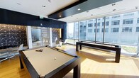 This contemporary amenity space is designed for recreation and relaxation, offering an impressive array of features within a sleek, modern interior. The centerpiece of the room is a full-sized pool table with a tan felt surface, positioned to take full advantage of the abundant natural light that streams in through the expansive floor-to-ceiling windows. Alongside it, a shuffleboard table adds another layer of entertainment to the space. The large windows frame sweeping views of the surrounding urban skyline, creating a bright and energizing atmosphere throughout the day. The flooring is a warm, light-toned hardwood that contrasts elegantly with the bold dark ceiling accents and architectural beams overhead, giving the space a polished, high-end aesthetic. Recessed lighting supplements the natural light, ensuring the space is well-illuminated at all hours. A separate lounge area is visible through glass partition doors, furnished with chairs and a table, offering a quieter retreat for conversation or casual meetings. A decorative patterned wall panel adds a stylish design element to the lounge nook. Together, these features create a well-rounded, sophisticated recreational environment suited for socializing and leisure.