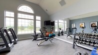 This fitness room is thoughtfully designed to support an active lifestyle, combining functional equipment with an open, airy atmosphere. The space is defined by its striking vaulted ceiling, which rises dramatically to create a sense of spaciousness and volume rarely found in exercise rooms. Large arched and rectangular windows line the walls, flooding the interior with natural light and offering pleasant outdoor views of surrounding greenery, enhancing the overall workout environment.

The flooring consists of neutral-toned carpet, providing comfort underfoot while maintaining a clean, cohesive aesthetic. The walls are painted in a soft gray-blue tone, contributing to a calm and focused atmosphere. A wall-mounted flat-screen television is positioned for easy viewing during workouts, accompanied by a media rack nearby.

Equipment includes stationary bikes, a weight bench, a treadmill, and stability balls in various sizes, offering a range of cardiovascular and strength training options. A large floor-to-ceiling mirror spans one wall, visually expanding the space while allowing users to monitor their form. Ample natural ventilation is supported by ceiling vents throughout the room.