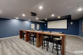 Located at 120 N Compass Way in Dania Beach, Florida, this purpose-built screening room delivers a true cinematic experience in a well-appointed, single-level layout. The space is defined by its bold navy-blue accent walls, a dark tray ceiling over the main viewing area, and light gray luxury vinyl plank flooring that runs throughout.

The primary seating area features multiple rows of plush leather recliner chairs arranged theater-style, each equipped with cup holders for added convenience. Directly facing the seating is a large, wall-mounted projection screen paired with a ceiling-mounted projector, creating an immersive viewing environment. Recessed lighting is thoughtfully positioned throughout the ceiling to provide ambient illumination without interfering with the viewing experience.

Along the rear of the room, a long wooden bar-height counter with black upholstered barstools offers an alternate seating arrangement, ideal for a more social viewing setting. Adjacent to this area, a compact kitchenette includes wood-toned cabinetry, a white countertop surface, a built-in microwave, and a mini refrigerator — making the space fully self-sufficient for entertaining. A floating wooden shelf above the kitchenette adds functional storage. Abstract wall art and clearly marked emergency exit signage complete the room's polished, amenity-ready presentation.