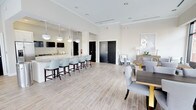 This single-floor space is designed with a clean, contemporary aesthetic that emphasizes openness and functionality. The layout seamlessly integrates a kitchen bar area and a dining space, creating a versatile environment suited for both casual gatherings and more structured meetings or events.

The kitchen bar runs along one side of the room, featuring a sleek white countertop lined with light blue upholstered counter-height chairs. Above the bar, a series of pendant lights with white shades provide focused illumination, complementing the recessed lighting throughout the ceiling. Wall-mounted flat-screen televisions are positioned above the bar area, adding a modern, lounge-like quality. The cabinetry is finished in white, maintaining the room's cohesive neutral tone.

The dining area occupies the opposite side of the space, furnished with dark rectangular tables paired with plush gray upholstered chairs. Gold-accented table bases introduce a refined decorative touch. The flooring throughout consists of light wood-look tile laid in a linear pattern, contributing to the airy, bright atmosphere. Crown molding frames the elevated ceilings, adding a polished architectural detail. Black interior doors provide a striking contrast against the predominantly white walls, grounding the overall design.