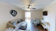 This single-floor space offers a thoughtfully designed interior with a relaxed coastal aesthetic. The living area serves as the heart of the space, showcasing rich hardwood-style flooring that contrasts beautifully with the light, neutral-toned walls. Crown molding lines the ceiling throughout, adding a refined architectural detail that elevates the overall ambiance. A classic five-blade ceiling fan with a built-in light fixture provides both comfort and charm.

Natural light pours in through a full-length glass door that opens to an exterior view, creating an airy and welcoming atmosphere. The living area is furnished with a plush, light gray sofa accented with blue and floral throw pillows, complemented by a patterned accent chair and a tufted ottoman in matching coastal tones. Decorative elements — including a circular wall art piece and tasteful tabletop accessories in shades of blue — reinforce the cohesive design theme.

A sleek flat-screen television is mounted on the wall, while a white console table provides functional surface space. The overall ambiance is bright, comfortable, and well-coordinated, making this space both inviting and practical for everyday living.