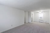 This single-floor residence offers a practical and well-organized layout spread across several connected rooms. The living area is generously proportioned, featuring neutral-toned wall-to-wall carpeting, crisp white walls, and a ceiling fan with a light fixture that serves as the central overhead illumination. Natural light filters in through windows fitted with blinds, and a wall-mounted radiator unit provides heating. The open sightlines from the living area extend into a hallway that connects the remaining rooms, creating a cohesive flow throughout the space.

The kitchen is compact yet functional, equipped with light wood cabinetry, dark countertops, a double-basin stainless steel sink, and a white gas range. A window above the counter area allows natural light in and offers an outdoor view. The tile flooring in the kitchen provides a practical transition from the carpeted living spaces.

The bedroom mirrors the living area in finish, featuring the same neutral carpeting, ceiling fan, radiator heating, and a window that brings in natural light. A closet is accessible from within the room.

The bathroom is finished with classic white subway tile on the walls, a distinctive checkered mosaic tile floor, a full bathtub, toilet, vanity, and a traditional cast-iron radiator positioned beneath a frosted window, contributing to a timeless, functional aesthetic.