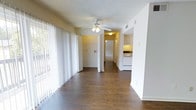 This single-floor residence offers a clean, functional layout with a welcoming open-plan living area at its heart. Rich, dark hardwood-style flooring runs throughout, providing a warm contrast to the bright white walls and textured ceiling. A ceiling fan with integrated lighting anchors the main living space, which is bathed in natural light streaming through expansive floor-to-ceiling vertical blinds that line an entire wall — offering both privacy and a seamless connection to the outdoors.

The open layout flows naturally toward the interior of the home, where a hallway leads to additional rooms. The kitchen is visible from the main living area and features white cabinetry, white appliances, and under-cabinet lighting, creating a bright and practical cooking space. The adjacent wall includes a pass-through or utility niche, adding to the functional design of the layout.

The neutral color palette of soft whites and warm yellows throughout gives the space an airy, inviting ambiance. The straightforward, open design makes the space feel connected and easy to navigate, with clear sightlines from the living area through to the kitchen and hallway beyond. Overall, this single-level residence combines practicality with a light, comfortable atmosphere.