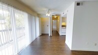 This single-floor residence offers a clean, functional layout with a welcoming open-plan living area at its heart. Rich, dark hardwood-style flooring runs throughout, providing a warm contrast to the bright white walls and textured ceiling. A ceiling fan with integrated lighting anchors the main living space, which is bathed in natural light streaming through expansive floor-to-ceiling vertical blinds that line an entire wall — offering both privacy and a seamless connection to the outdoors.

The open layout flows naturally toward the interior of the home, where a hallway leads to additional rooms. The kitchen is visible from the main living area and features white cabinetry, white appliances, and under-cabinet lighting, creating a bright and practical cooking space. The adjacent wall includes a pass-through or utility niche, adding to the functional design of the layout.

The neutral color palette of soft whites and warm yellows throughout gives the space an airy, inviting ambiance. The straightforward, open design makes the space feel connected and easy to navigate, with clear sightlines from the living area through to the kitchen and hallway beyond. Overall, this single-level residence combines practicality with a light, comfortable atmosphere.