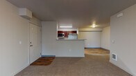Located at 352 North Beck Road in Post Falls, Idaho, this single-floor unit offers a functional and comfortable layout designed for everyday living. The open-concept living area is finished with plush neutral carpeting and features a glass patio door fitted with vertical blinds, allowing natural light to fill the space. A wall-mounted heating unit and multiple outlets make the living area practical and ready for furnishing.

The kitchen is positioned behind a half-wall peninsula that doubles as a breakfast bar, creating a natural separation between the living and cooking spaces. Rich dark-wood cabinetry pairs with light marbled countertops, and the kitchen is equipped with stainless steel appliances including a refrigerator, over-the-range microwave, and a coil-top range. The kitchen floor features warm wood-look hard surface flooring, which continues into the hallway beyond.

The hallway connects the main living spaces to the private areas of the unit, with paneled doors leading to additional rooms and closet storage. A dedicated laundry area houses a full-size front-loading washer and dryer alongside a water heater, providing added convenience. The overall aesthetic throughout the unit is clean and neutral, with light gray walls, white trim, and a consistent use of warm-toned flooring and cabinetry that creates a cohesive, welcoming ambiance.