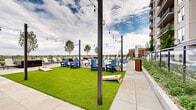 Located at 3444 Emerson St in Englewood, Colorado, this generously sized rooftop amenity deck offers a thoughtfully designed outdoor retreat that combines recreation, relaxation, and striking open-sky views of the surrounding Colorado landscape. The space is anchored by a sparkling resort-style swimming pool with an adjoining spa, both finished in crisp tile and surrounded by expansive large-format concrete pavers. Abundant lounge seating — including rows of reclining chaise lounges and clusters of Adirondack chairs in bold blue — is arranged throughout, providing ample room for sunbathing or socializing. A lush artificial turf lawn adds a vibrant pop of greenery and serves as a casual gathering and games area, complete with cornhole boards and strung Edison-style globe lights overhead that lend a festive evening ambiance. Raised planter beds filled with ornamental grasses, native plantings, and young trees introduce natural texture and soften the hardscape throughout. Fire pit stations and additional seating areas create intimate gathering spots. A clean, modern walkway with glass-panel railings traces the perimeter of the deck, framing panoramic views of the skyline and open horizon. The overall design balances contemporary aesthetics with functional outdoor living, making it a standout communal amenity.