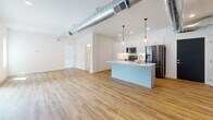 This modern single-floor unit combines clean contemporary design with industrial-inspired elements to create a bright, open living environment. The expansive open-concept living and kitchen area is anchored by warm hardwood-style flooring and striking exposed silver ductwork that runs the length of the ceiling, lending the space a distinctive urban character. Recessed lighting and stylish pendant lights illuminate the area throughout.

The kitchen is well-appointed with white flat-panel cabinetry, a subway tile backsplash, stainless steel appliances including a refrigerator with ice maker, electric range, and over-the-range microwave, and a generous central island featuring a quartz countertop and an undermount sink with a brushed nickel faucet. Sliding glass doors in the living area open to a private balcony with outdoor views.

The bedroom offers plush carpeting, a large window with natural light and scenic views, and access to a spacious walk-in closet with built-in shelving and hanging rods. Two bathrooms serve the unit — one featuring a soaking tub with white subway tile surround and brushed brass fixtures, the other a well-organized vanity with white cabinetry and gold-toned hardware — both finished with light porcelain tile flooring for a clean, polished look.