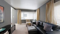 This efficiently designed one-bedroom, one-bathroom unit makes excellent use of its compact footprint, organizing distinct living, dining, kitchen, and sleeping spaces across a single floor. The living room serves as the centerpiece, featuring warm-toned carpet, bold grey walls, and multiple windows dressed with neutral blinds and floor-length gold drapes that allow generous natural light. A geometric mirror wall accent and large framed artwork add visual interest to the space.

The separate dining area features hardwood flooring, white walls, and a window with blinds, providing a bright and clean setting for everyday meals. Adjacent to it, the kitchen is appointed with white cabinetry, light laminate countertops, a stainless steel sink, and a coil-top range, offering functional essentials in a tidy layout.

The bedroom is a cozy, carpeted retreat with a window, neutral walls, and ample space for a full-sized bed. A hallway with angled architectural detailing connects the rooms and includes a decorative mirrored wall element, adding character to circulation space. The bathroom features a full tub-shower combination with white tile surround and chrome fixtures, completing this well-organized unit.