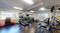 This fitness center offers a comprehensive workout environment designed to accommodate a wide range of exercise needs. The space features dark hardwood-style flooring throughout, accented by protective rubber mats beneath the equipment, and is illuminated by a combination of recessed lighting and flush-mount fluorescent fixtures set against a clean white ceiling with defined beam detailing.

The cardio section is well-appointed with treadmills and an elliptical machine, while the strength training area includes a multi-function cable machine, a weight bench, and a full dumbbell rack. Additional equipment includes a stationary bike, a rowing machine, a balance board, and a punching bag, catering to diverse training styles. Stability balls and a standing floor fan round out the amenities.

A large wall-mounted mirror along one side of the room visually expands the space and supports proper form during workouts. Multiple windows provide ample natural light and views of surrounding greenery, creating an open, energizing atmosphere. A small utility counter with a sink is tucked into one area, offering added convenience. The room is accessed through a keyed entry door, ensuring a secure, dedicated fitness environment.