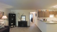 This single-floor space features an open, flowing layout that connects the living area to the kitchen and beyond. The living space is finished with wall-to-wall carpeting in a neutral tone, contributing to a warm and understated ambiance throughout. Smooth, light-colored walls and a flat ceiling enhance the sense of openness, allowing the space to feel airy and well-proportioned.

A partial kitchen is visible along one side, featuring wood-toned upper cabinetry and a countertop that extends as a dividing bar between the kitchen and the main living area. This layout creates a natural separation while maintaining visual connectivity between the two spaces.

A hallway leads from the main living area into additional rooms beyond, suggesting a practical and navigable floor plan. The corridor is carpeted and well-lit, providing a glimpse into a brighter adjoining room. A round wall mirror and decorative accents add visual interest without overwhelming the clean, neutral palette. Overall, the space presents a functional and comfortable single-level layout with consistent finishes and a cohesive, modest design aesthetic.