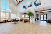 This expansive single-floor amenity space is thoughtfully designed to blend modern sophistication with warm, inviting character. The centerpiece of the space is a dramatic great room boasting soaring vaulted ceilings accented by rich exposed wood beams and clerestory windows that flood the interior with abundant natural light. Large-scale abstract artwork adorns the upper walls, adding a bold artistic statement to the open volume. Light-toned herringbone tile flooring anchors the main gathering area, which features generous lounge seating, a caramel-colored banquette, and lush indoor greenery including a striking potted tree that brings an organic element indoors.

The living room area transitions seamlessly into a cozy lounge with a textured tile feature wall, a mounted flat-screen display, and plush seating arrangements centered around a glass coffee table. Exposed wood beams continue throughout, maintaining design continuity. A well-appointed dining area offers an intimate setting highlighted by a sculptural gold pendant light and glass-paneled doors.

The gourmet kitchen is fully equipped with dark wood cabinetry, expansive light quartz countertops, stainless steel appliances including a large refrigerator, a deep sink, and pendant lighting. Large windows frame outdoor views, enhancing the bright and functional workspace. A wide communal island with upholstered seating completes the space, ideal for gatherings and collaborative use.